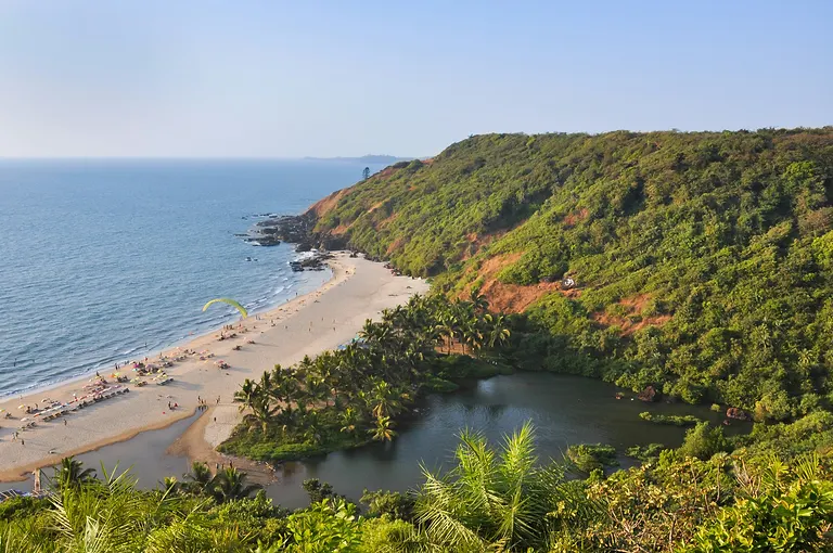 A curved Goa beach seen from the headland above — pale sand, beach umbrellas, a paraglider over the surf and a palm-ringed lagoon under the green cliff
